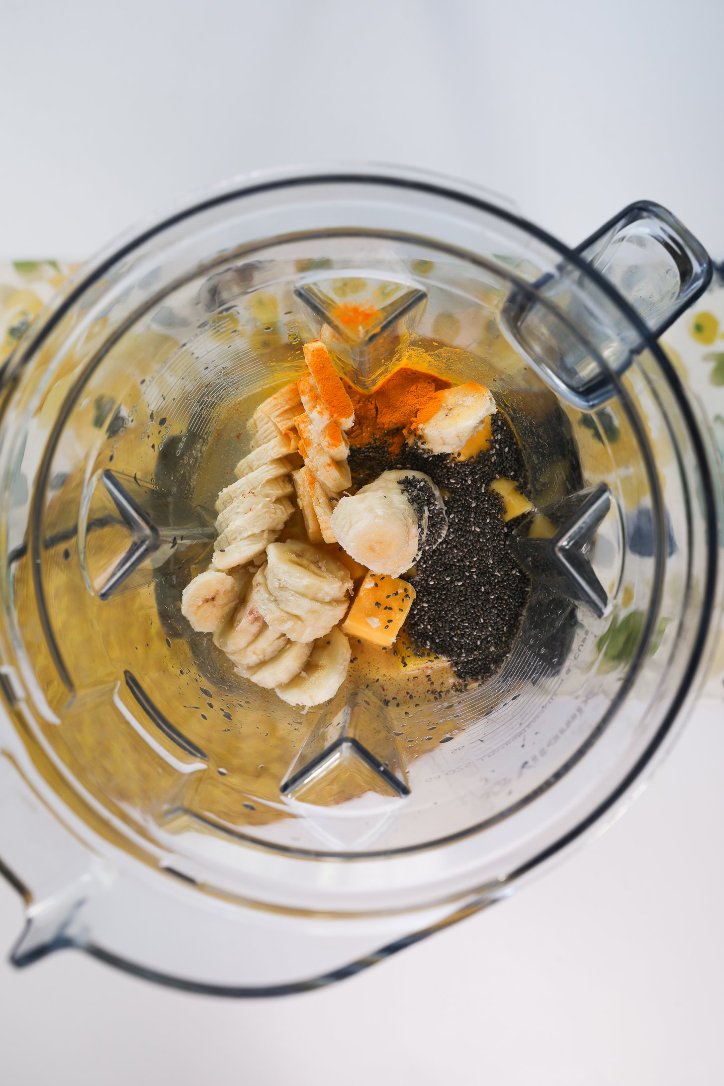 Top view of a blender filled with banana slices, mango and pineapple chunks, chia seeds and turmeric powder.