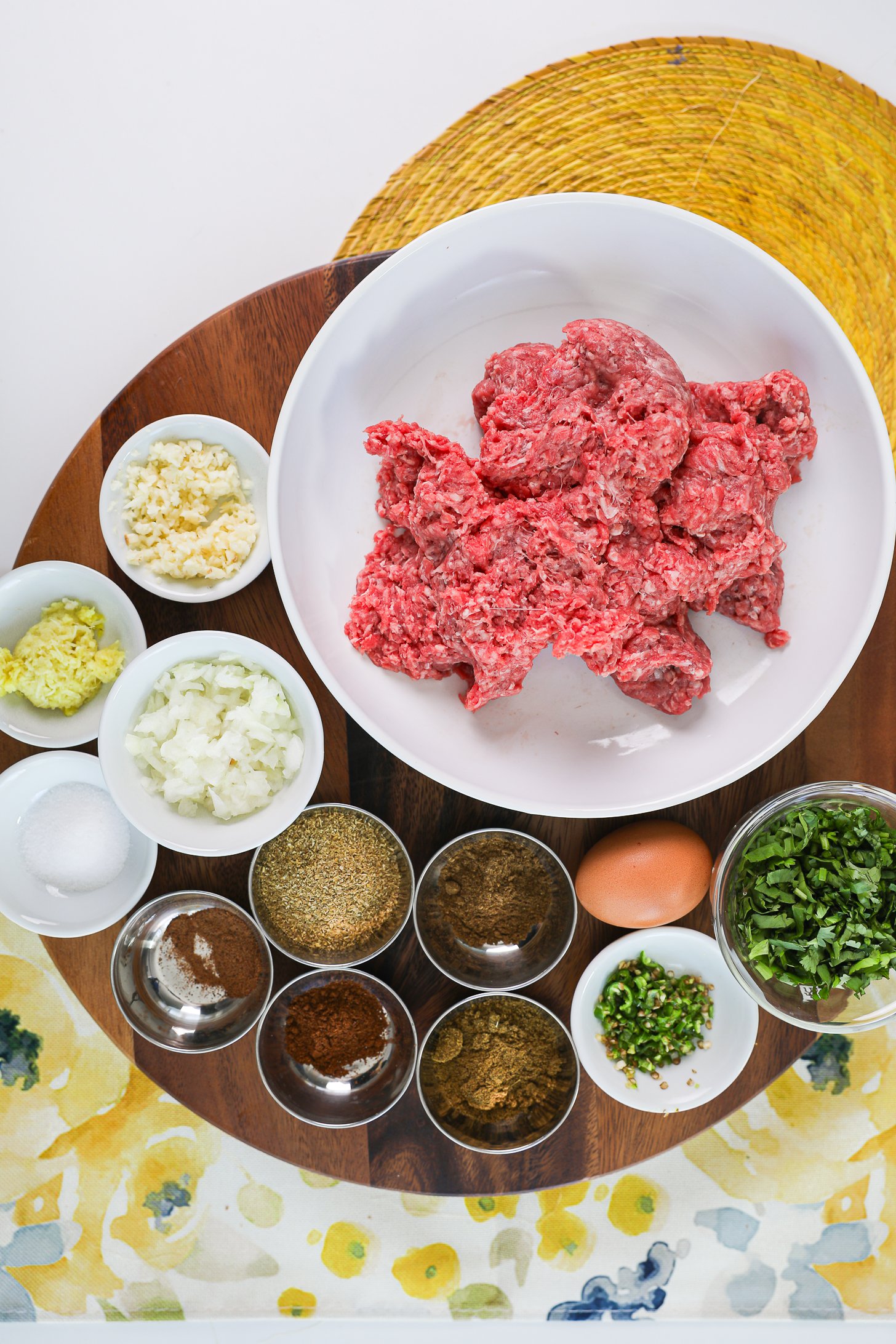 A selection of food ingredients including raw minced beef, ramekins of powdered spices, aromatics and herbs.