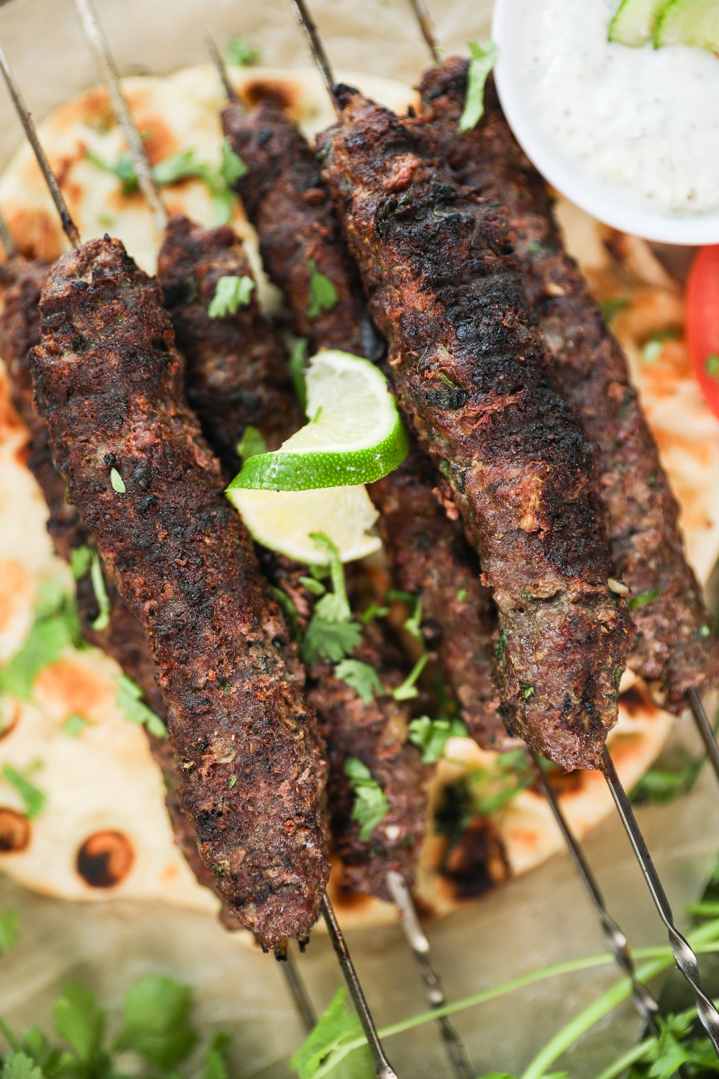Close-up top-down view of beef kebabs on skewers atop a naan, decorated with a lime swirl and cilantro leaves.