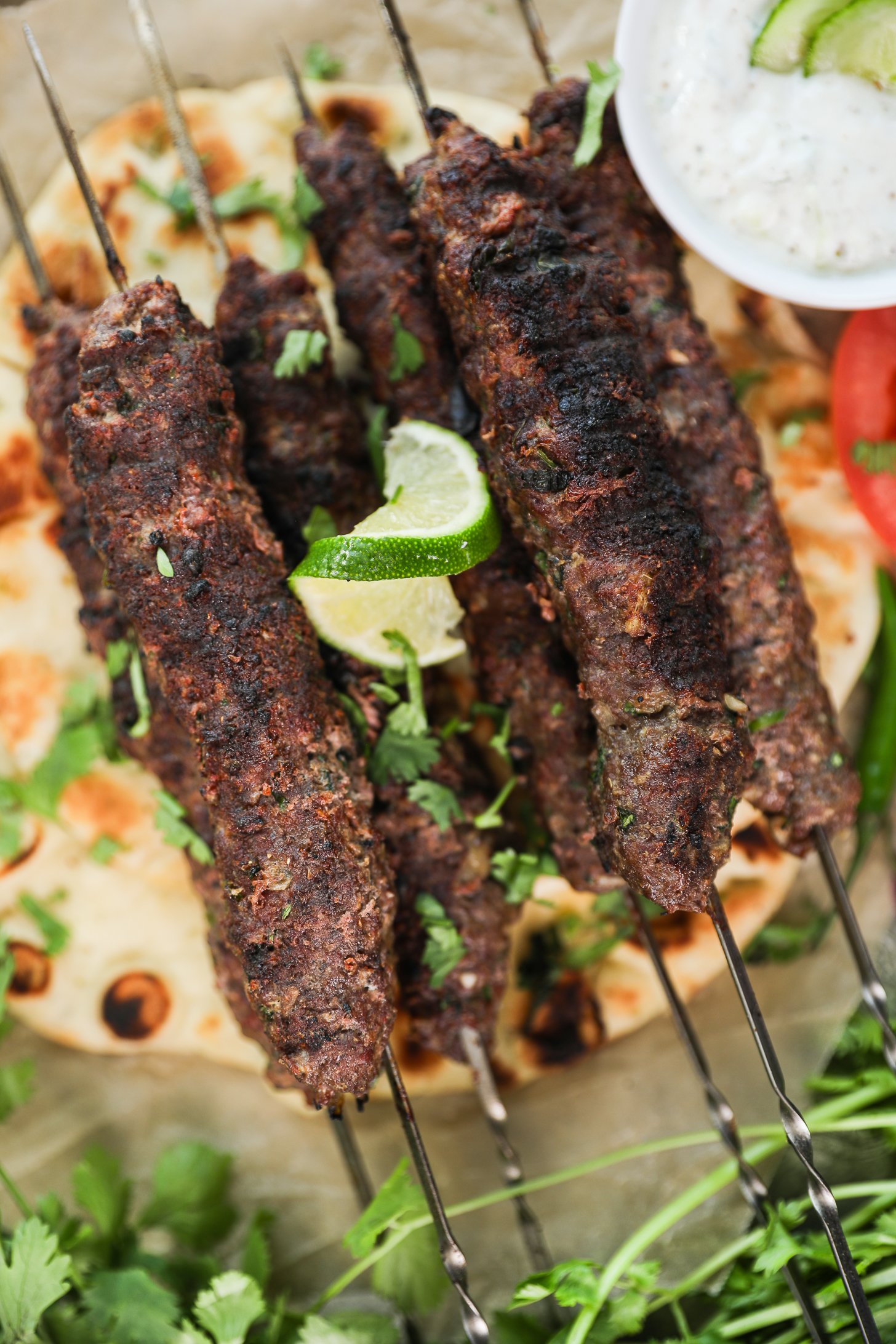 Close-up top-down view of beef kebabs on skewers atop a naan, decorated with a lime swirl and cilantro leaves and a ramekin of raita.