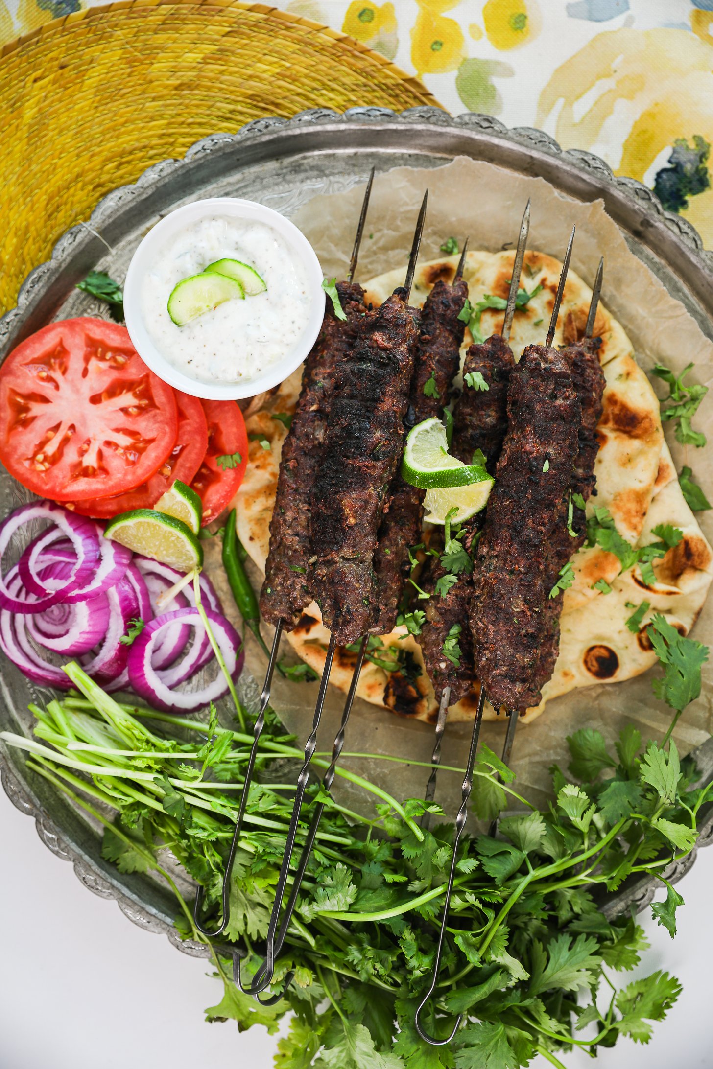Top-down view of grilled beef kebabs on skewers atop a naan, decorated on a large round tray with tomato rounds, onion rings, raita and a bunch of fresh cilantro.