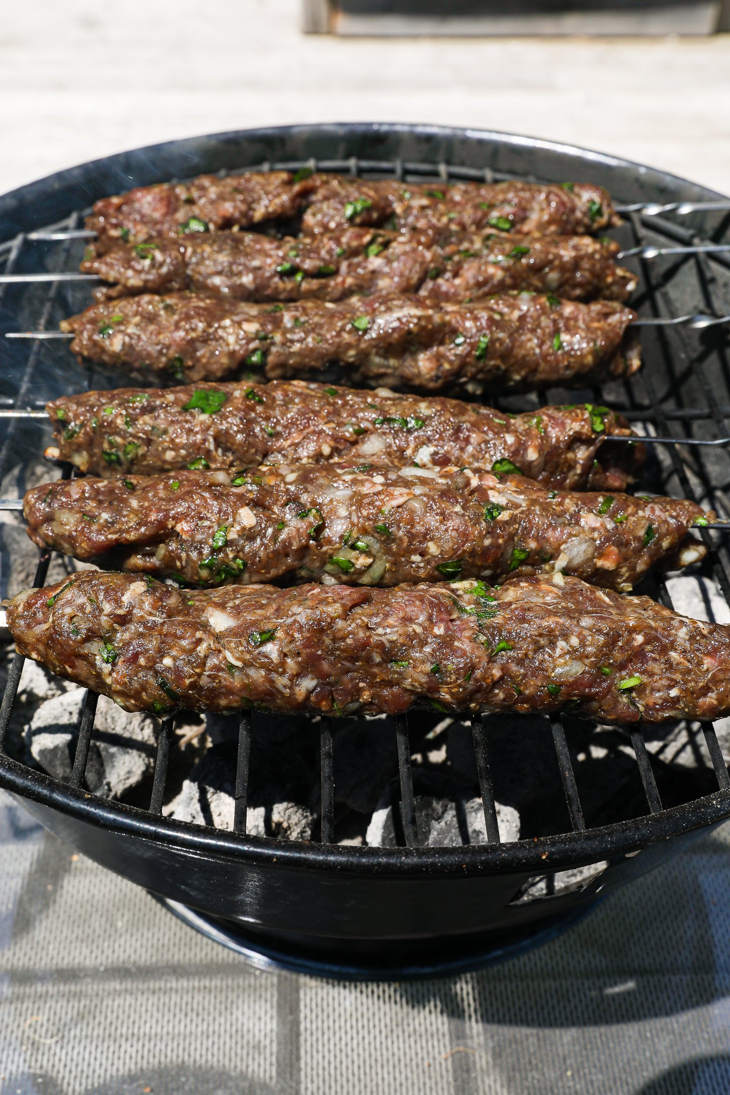 Perspective view of raw mince beef kebabs on skewers on an outdoor charcoal grill.