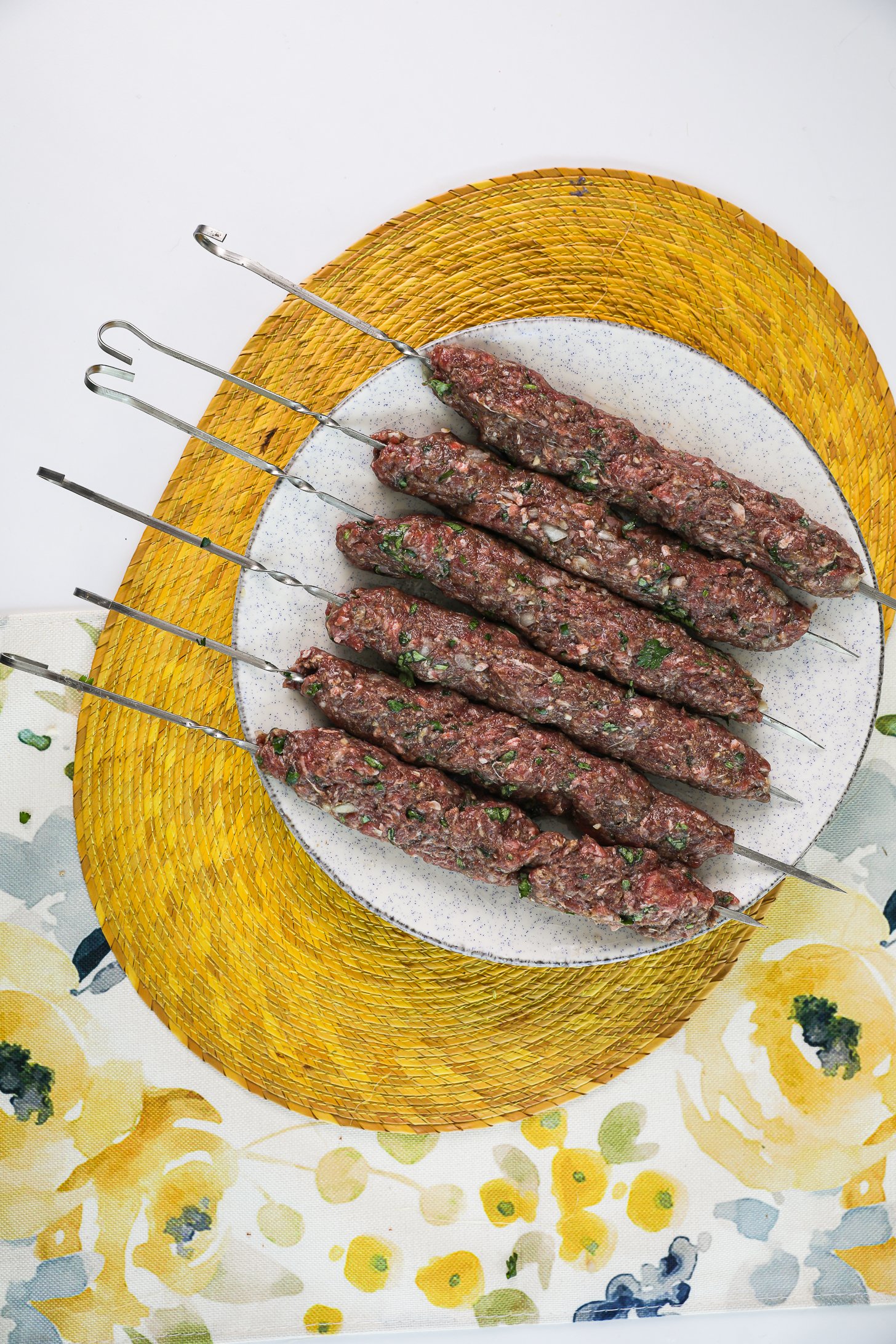 Perspective view of raw mince beef kebabs on skewers on a plate.