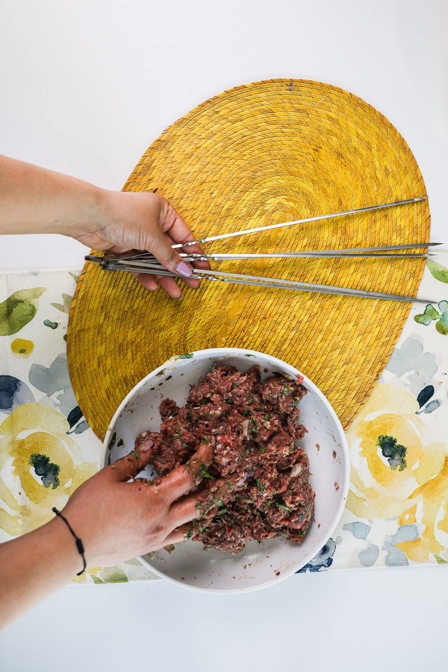 One hand submerged in a bowl of raw minced beef and the other hand holding metal skewers.