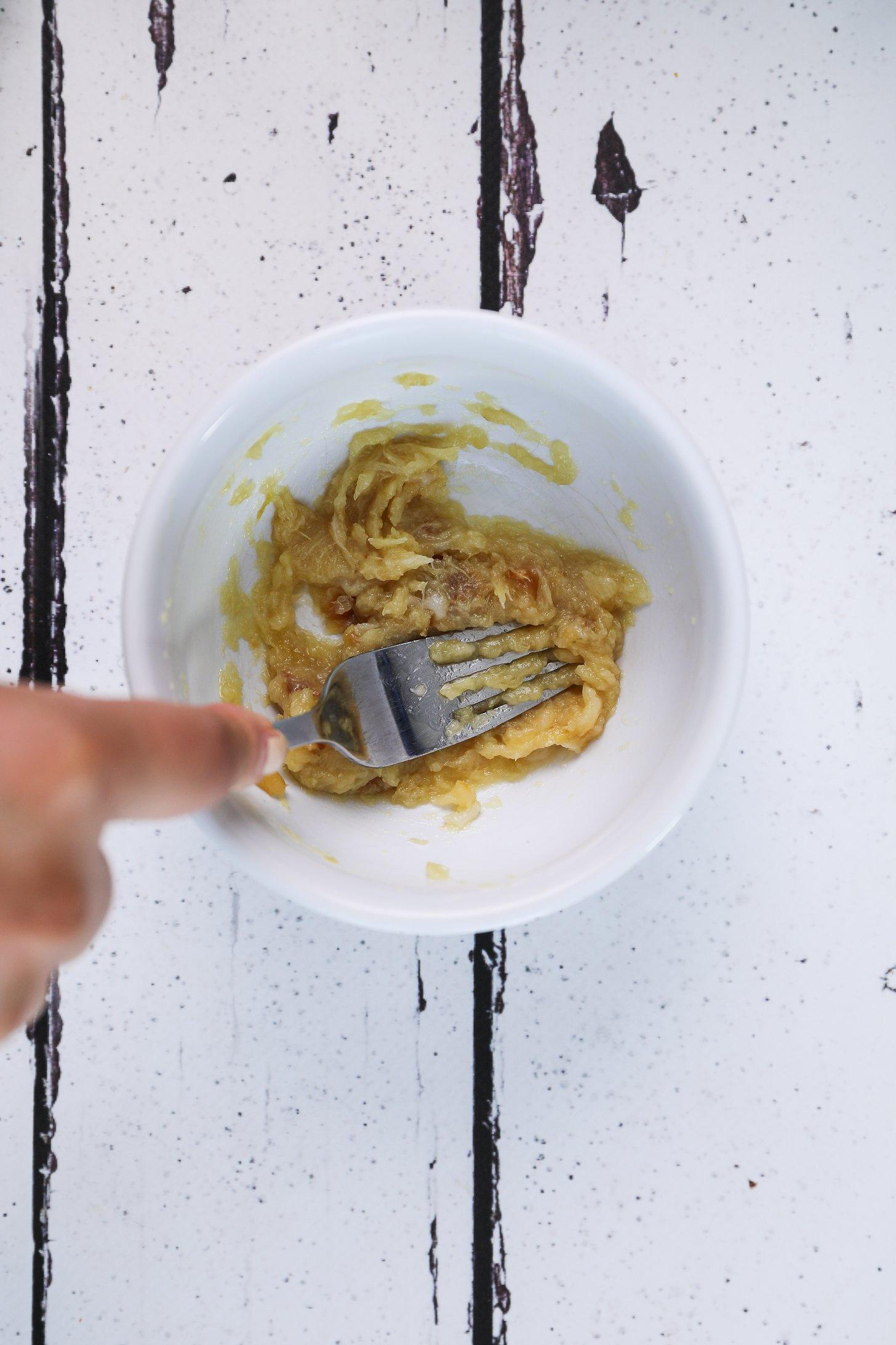 A hand holding a fork mashing soft roasted garlic in a bowl.