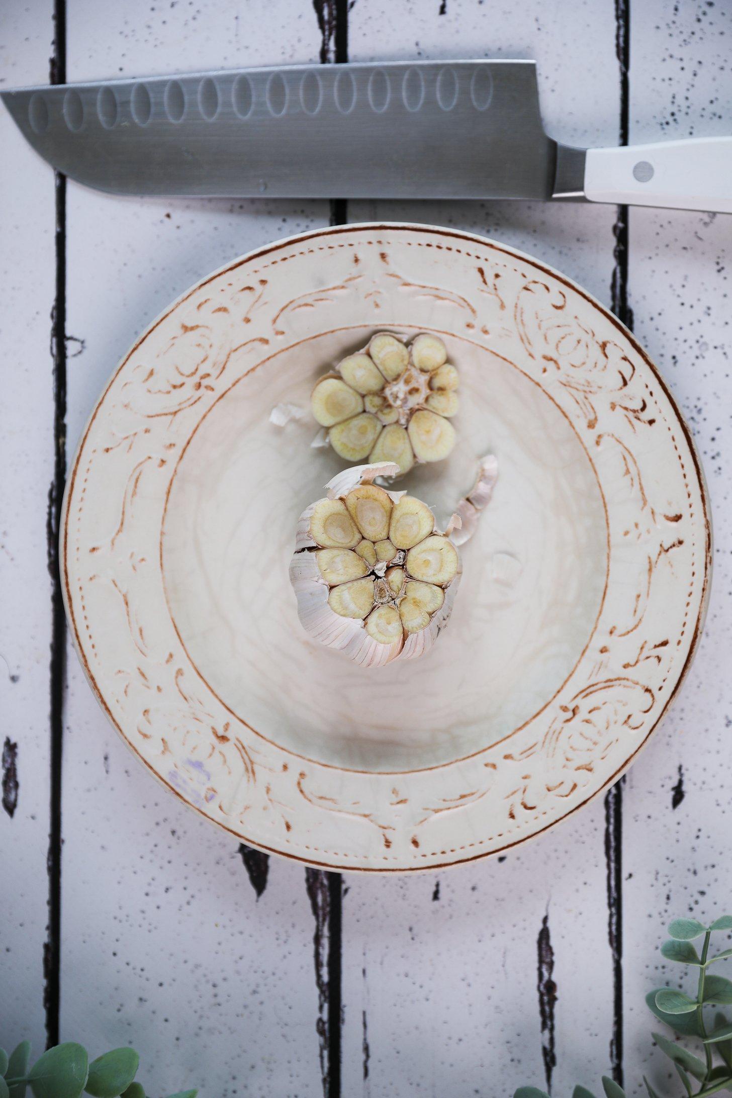 Overhead image of a garlic bulb with the top sliced off on a small plate and a knife close by.