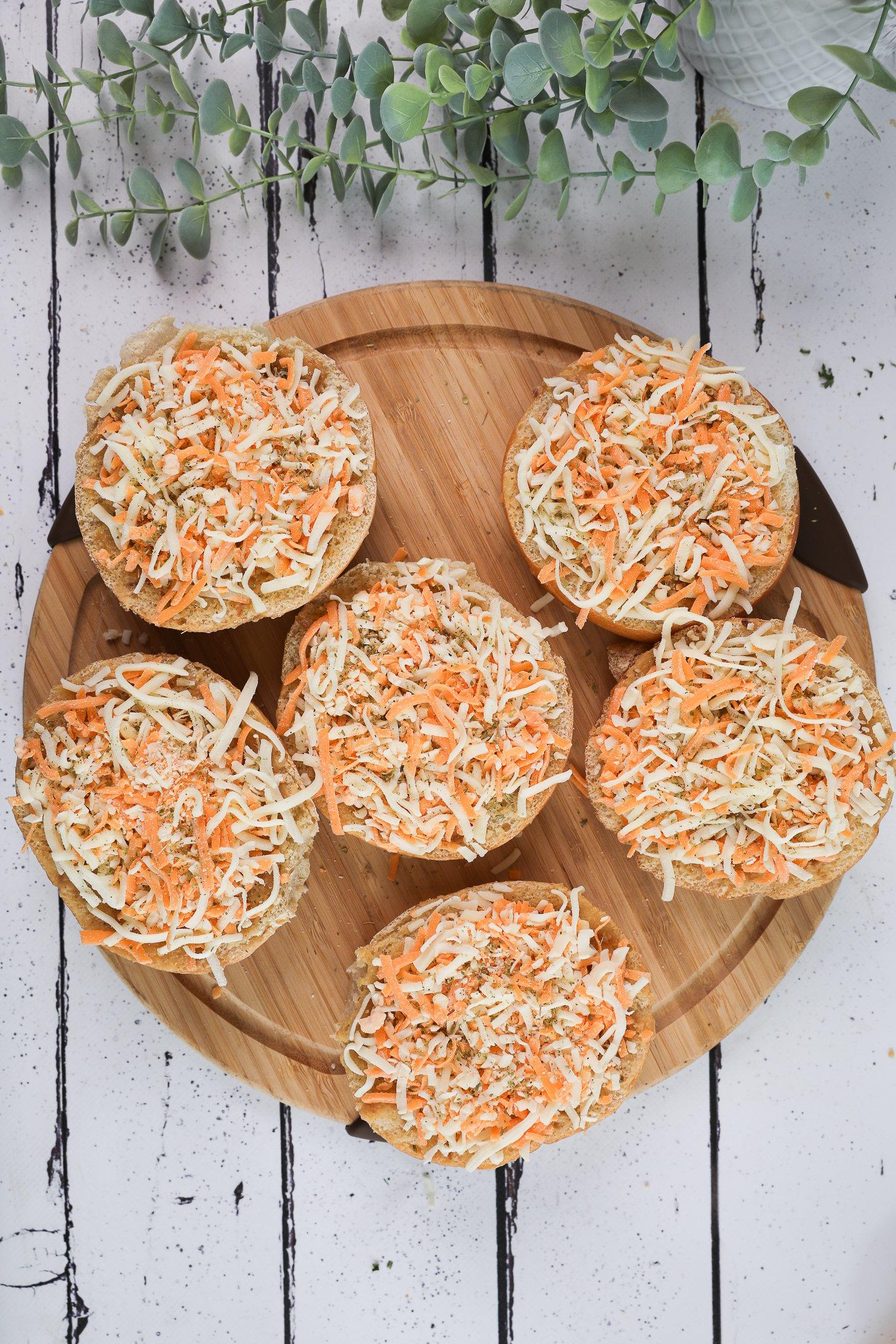 Birdseye image of of six round breads topped with grated cheese on a wooden board with a plant nearby.