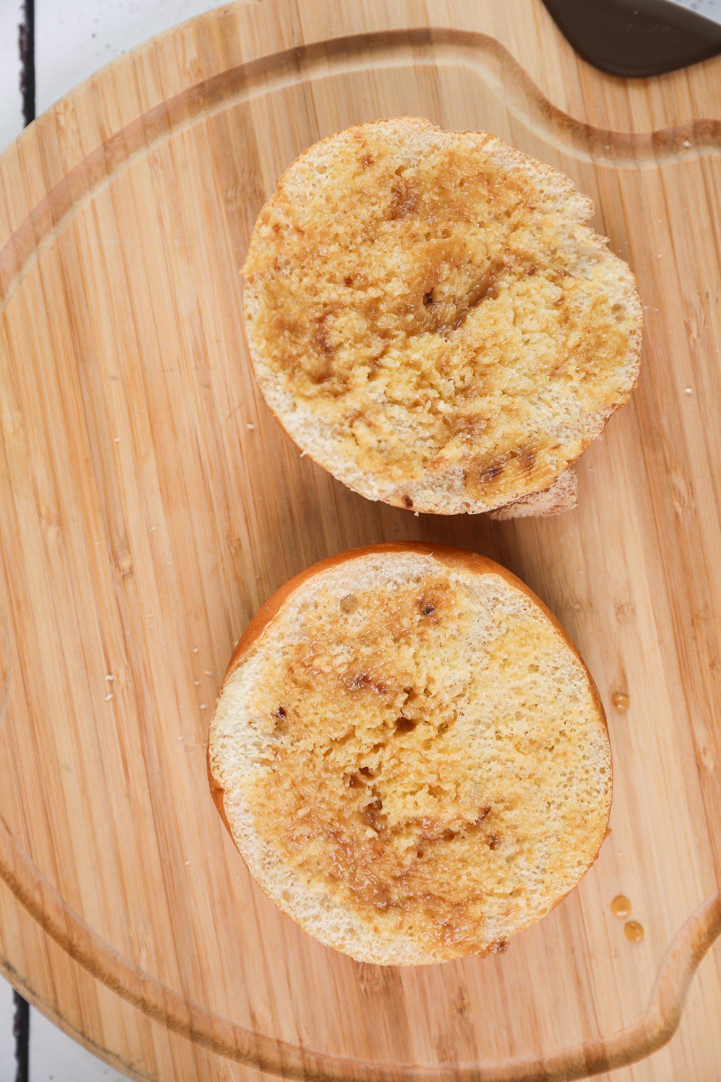 Two halves of a round Kaiser roll spread with roasted garlic butter on a wooden board.