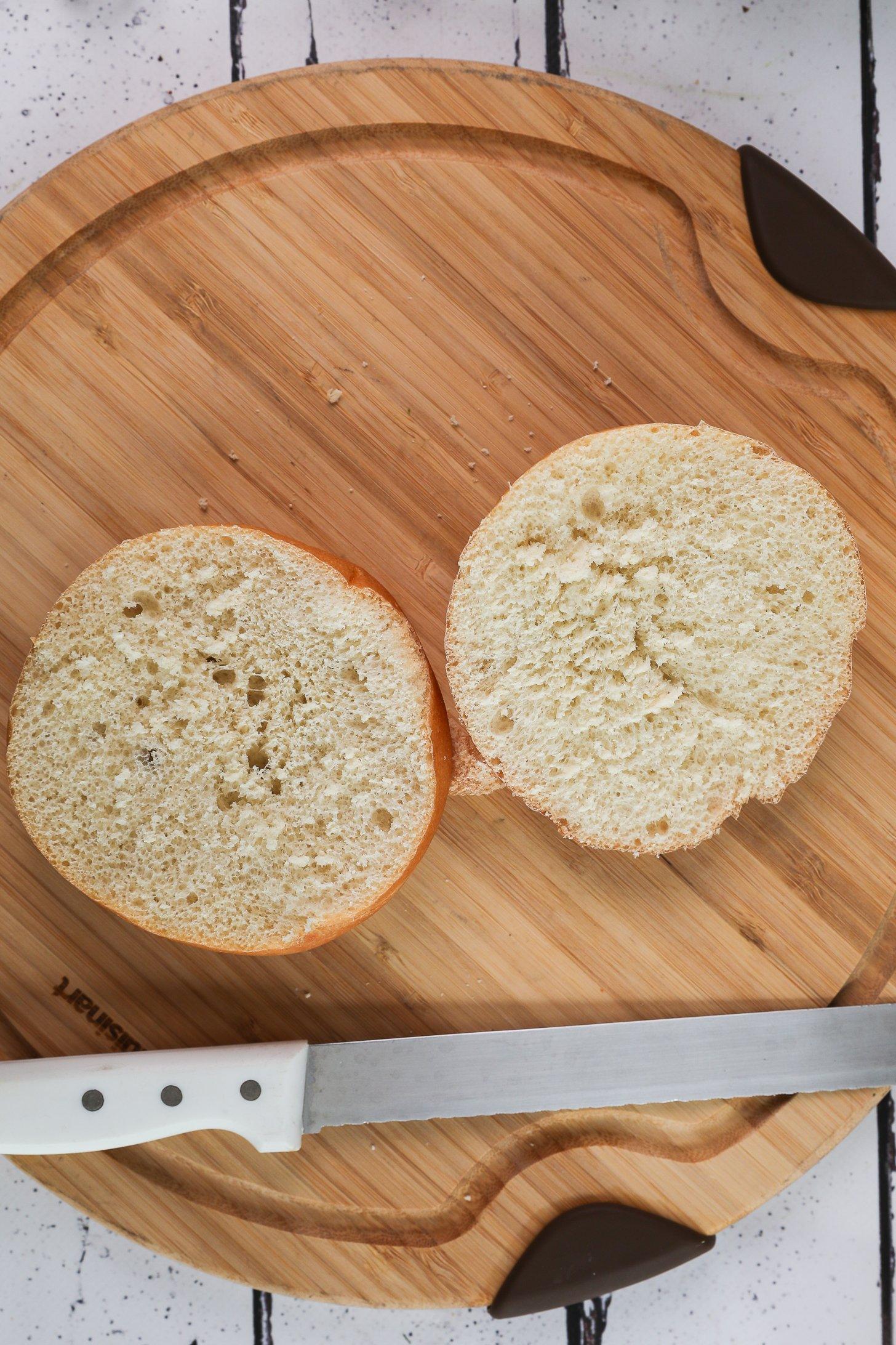Two halves of a round Kaiser roll on a wooden board with a bread knife nearby.