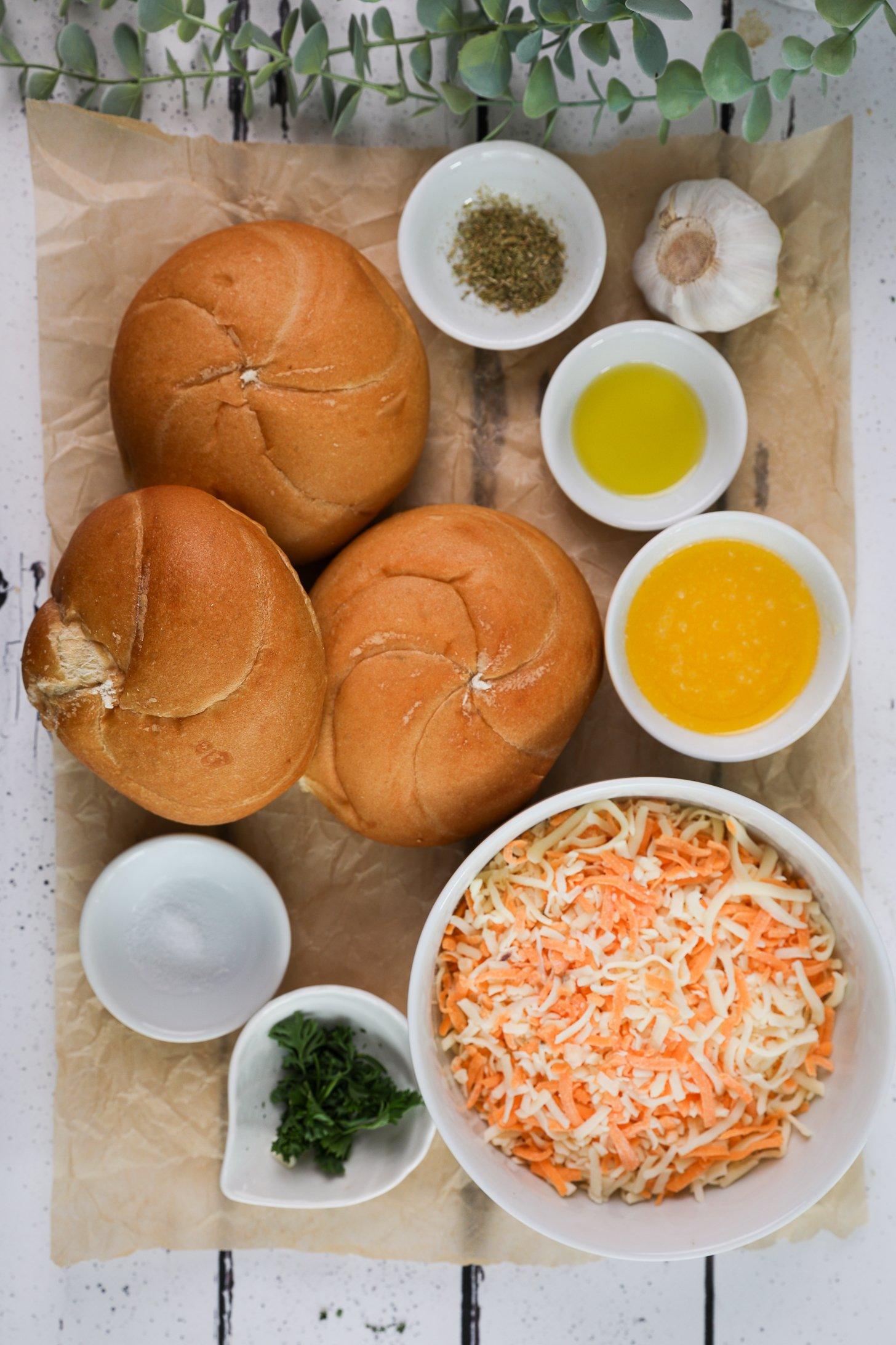 A selection of food ingredients including Kaiser bread rolls, melted garlic, oil, grated cheese, and parsley.