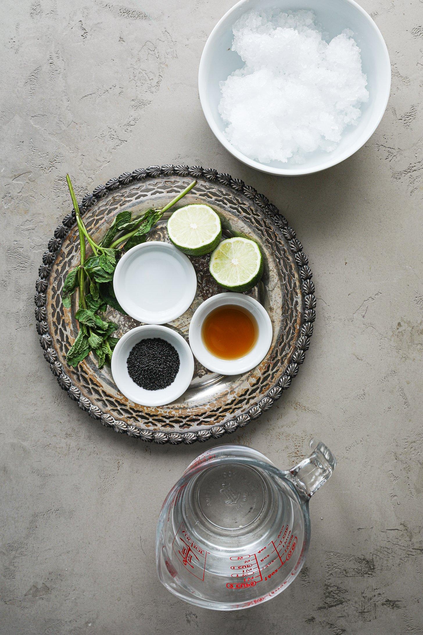 An image of food ingredients including maple syrup, lime, rose water and mint leaves styled on a silver tray with a bowl of crushed ice and a jug of water nearby.