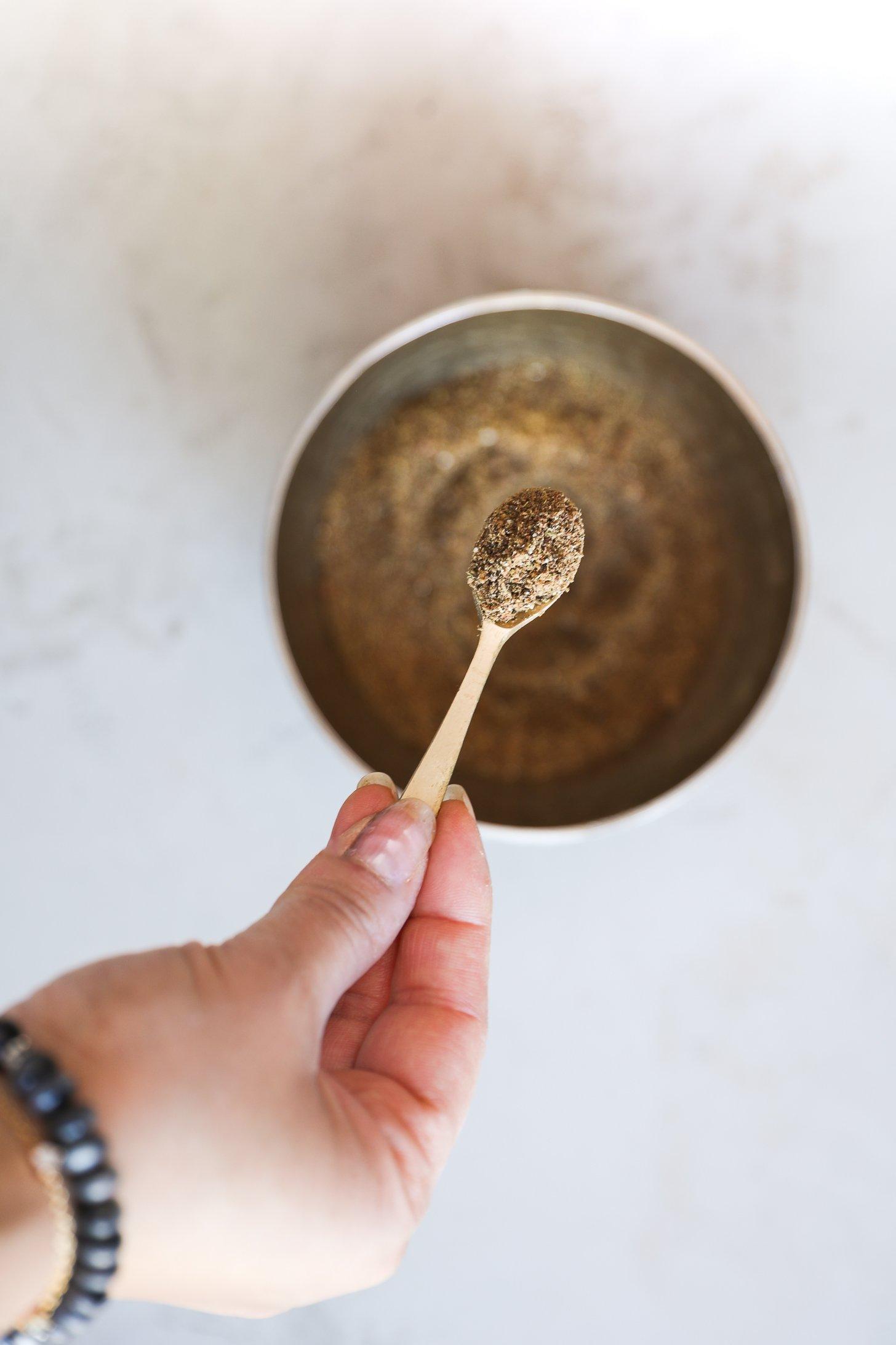 A hand holds a small spoon of freshly ground chai masala over a bowl.