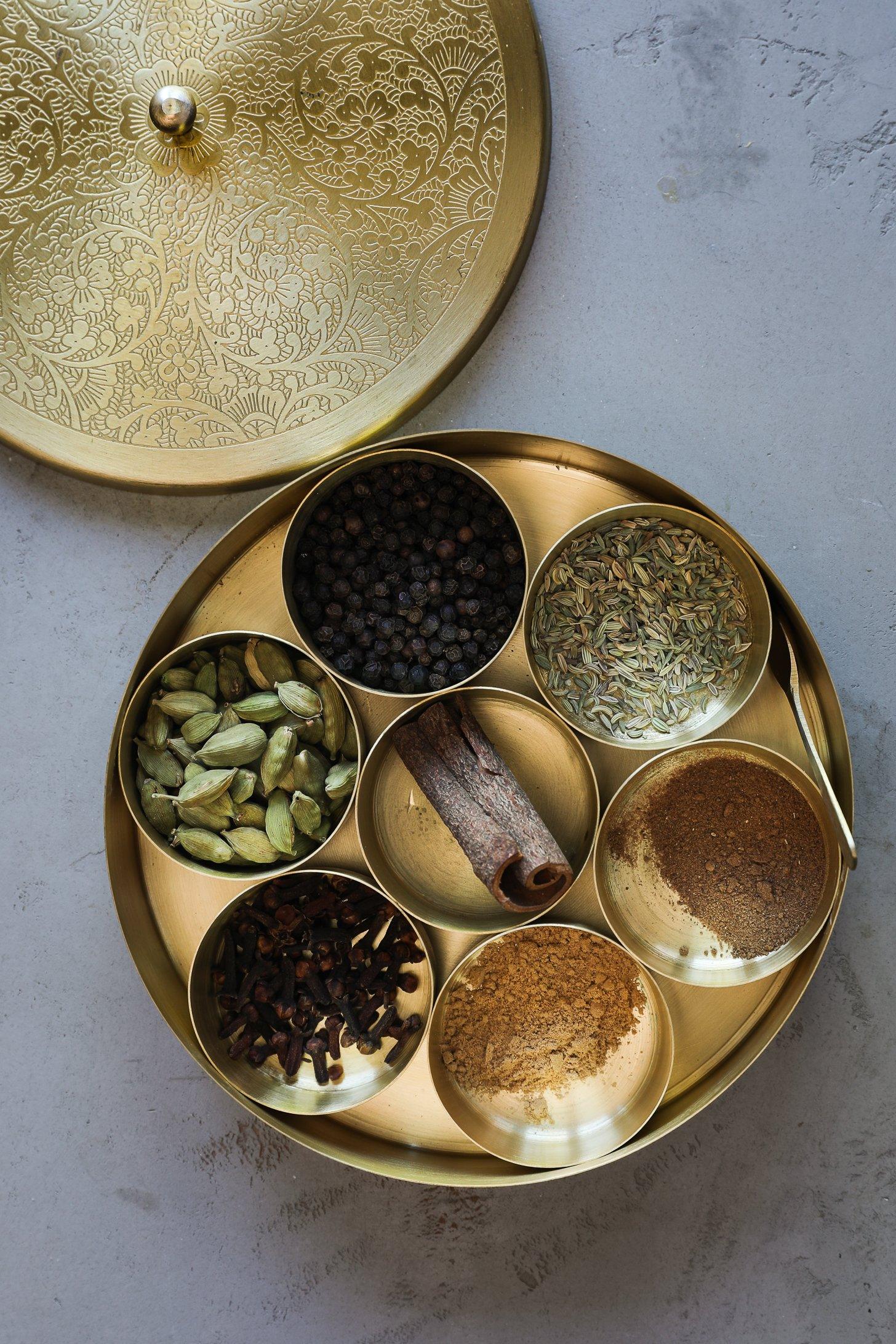 An open Indian spice box (masala dabba) filled with whole chai spices like cardamom, cinnamon, cloves, and fennel.