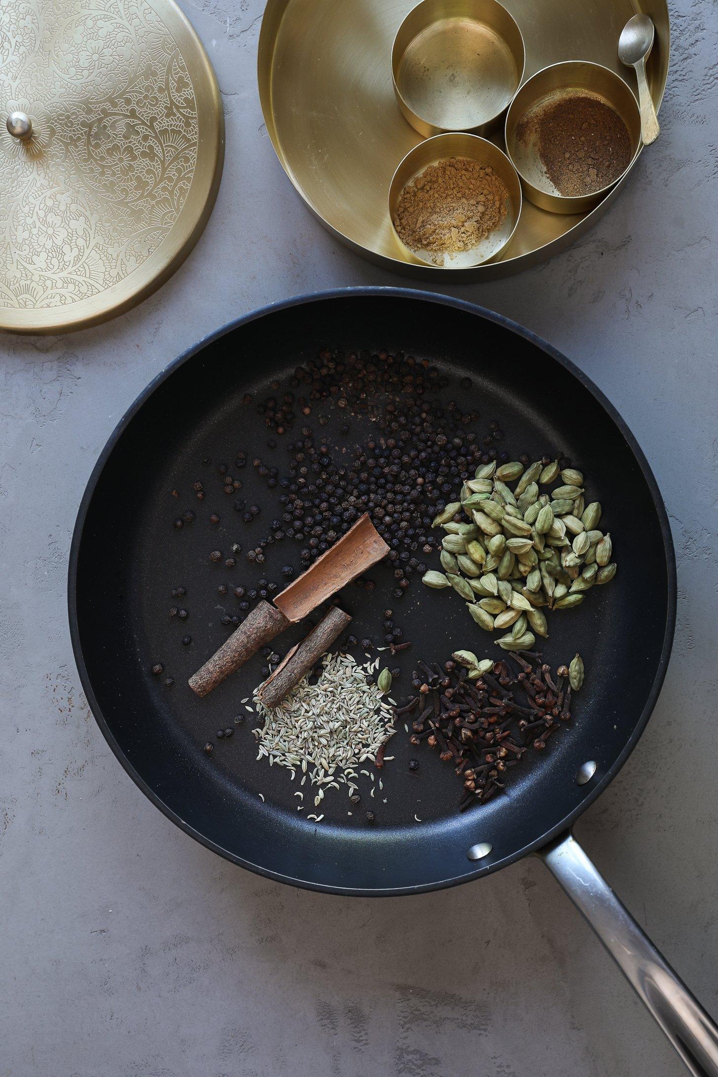 Whole chai spices like cardamom, black pepper, and cinnamon in a pan, ready for toasting.