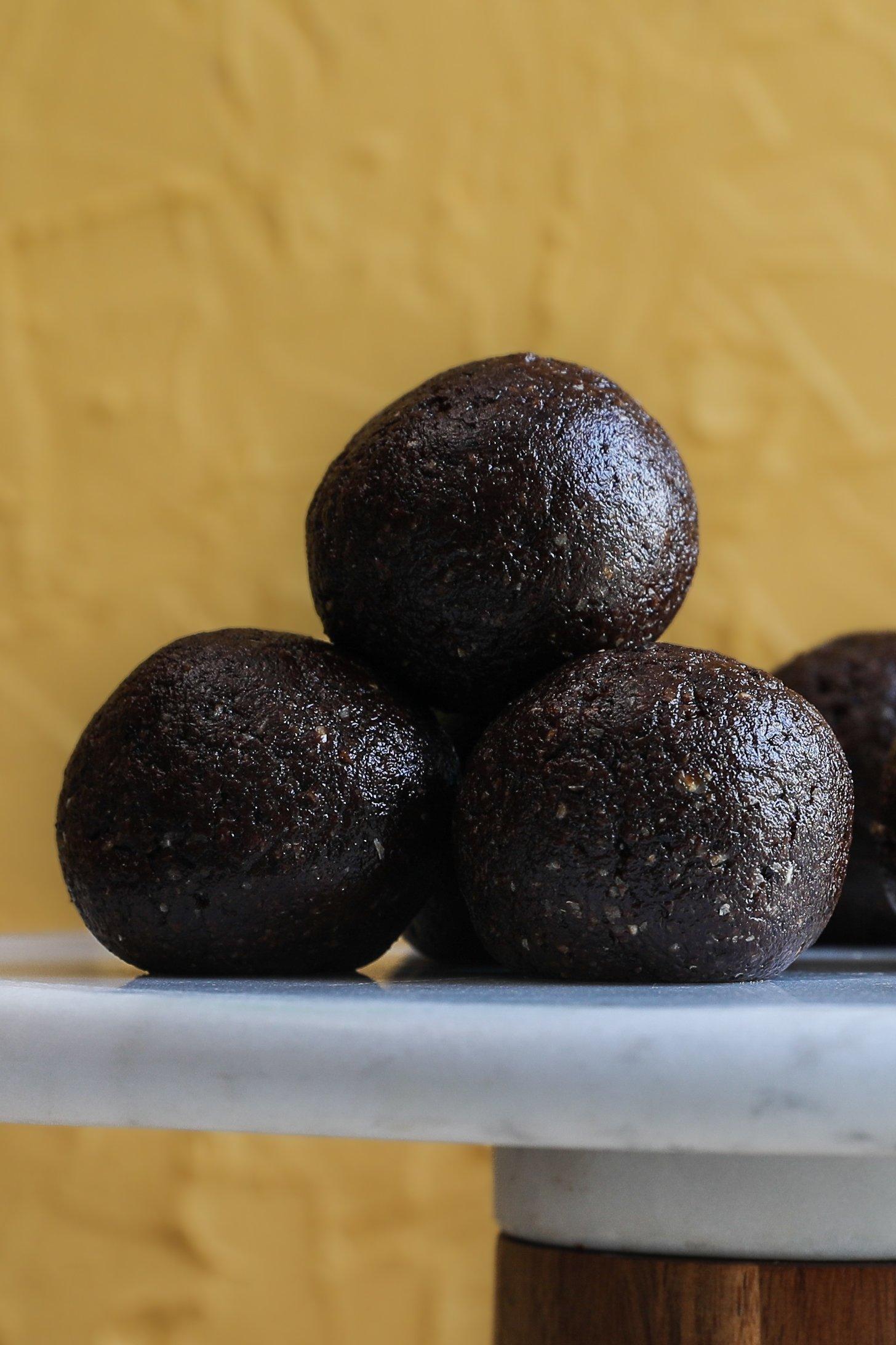 Close-up of three chocolate coconut ladoos stacked on a marble and wood pedestal, against a golden yellow background.