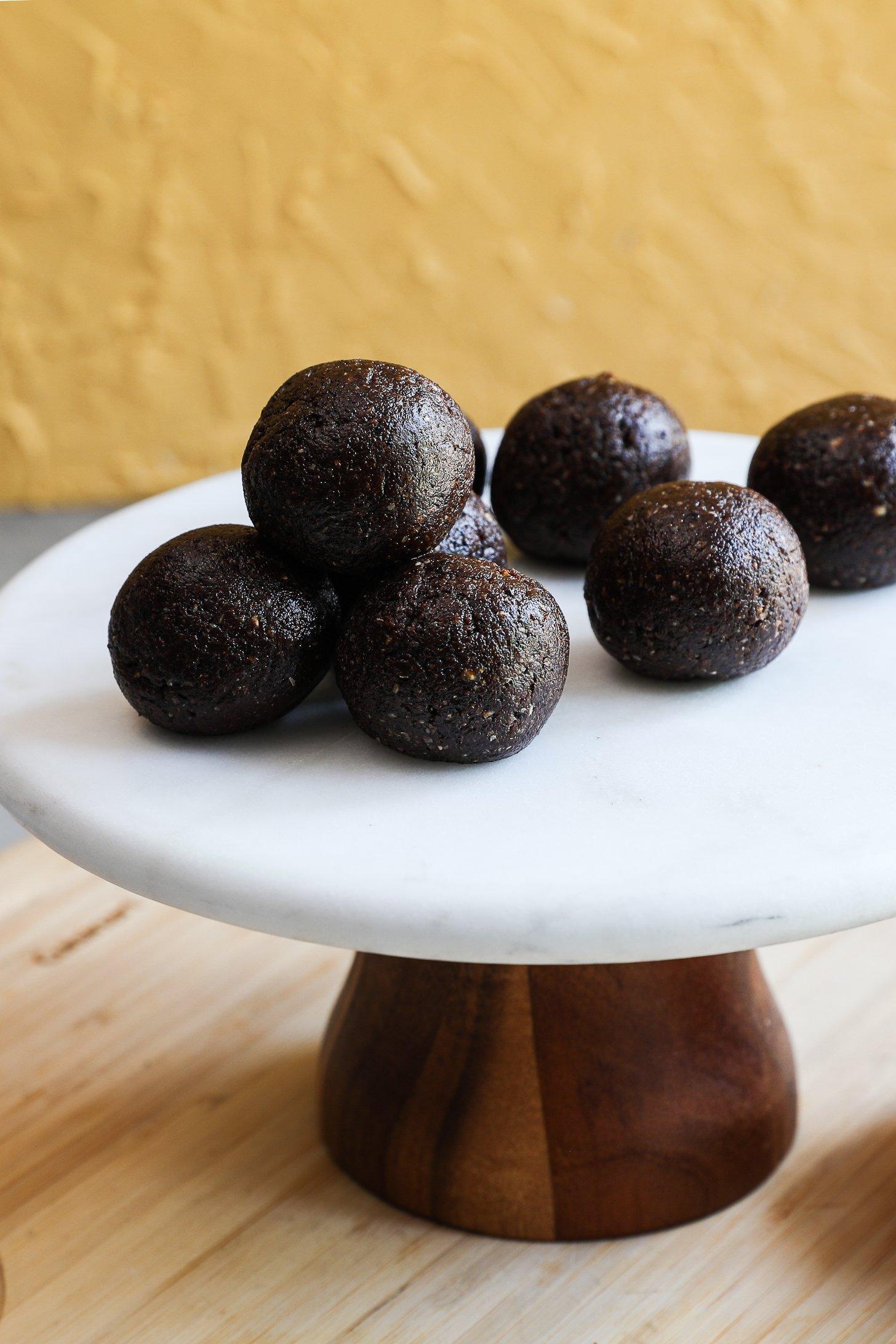 Six smooth and glossy chocolate coconut ladoos arranged on a white marble cake stand.