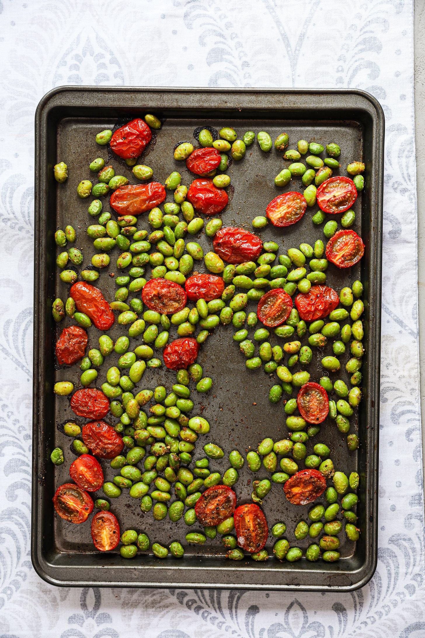 Roasted edamame and tomatoes spread on a dark baking tray.