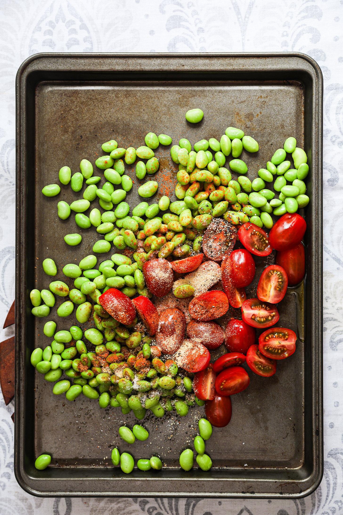 Raw edamame and halved cherry tomatoes tossed with spices on a baking tray.