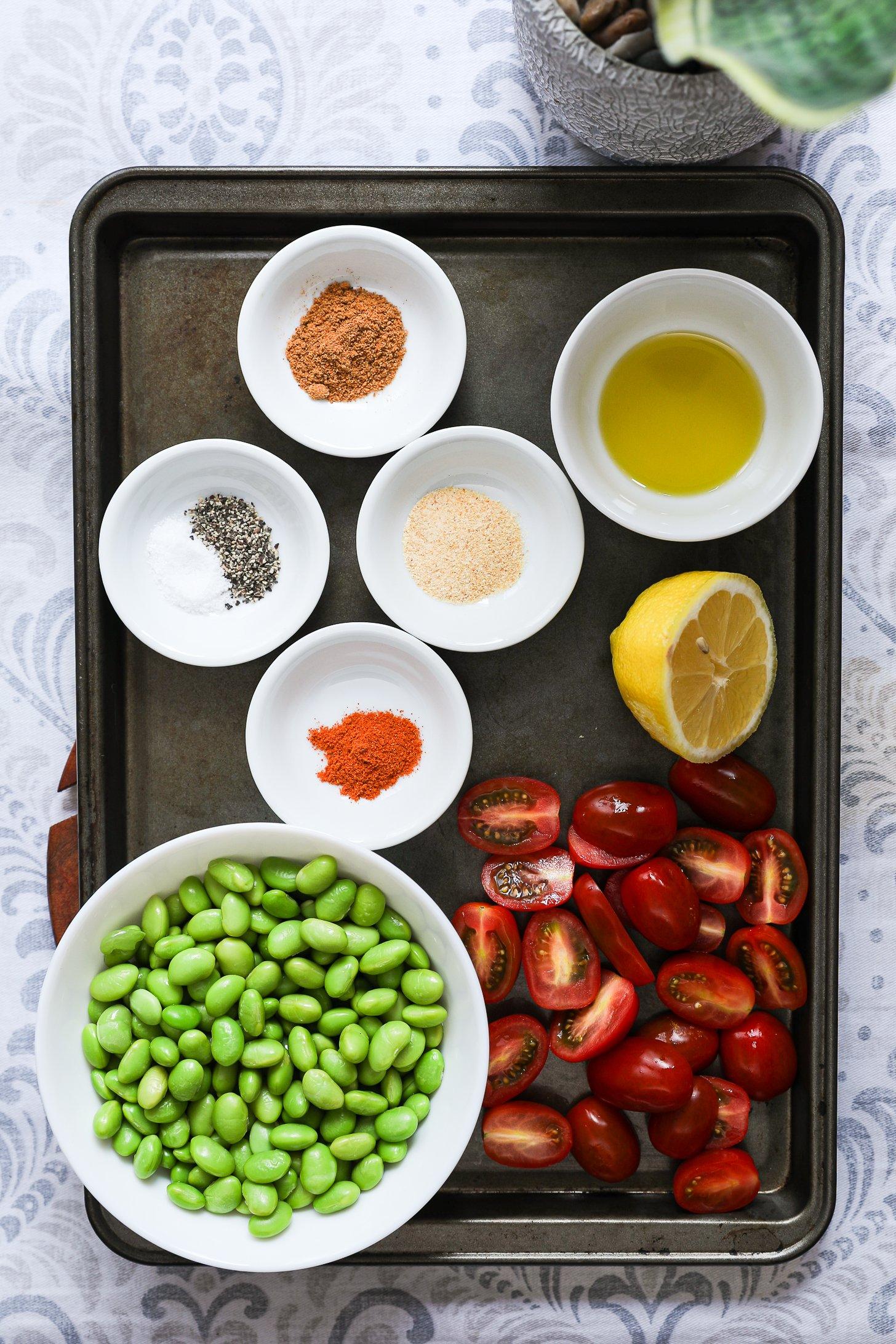 Flat lay of edamame, cherry tomatoes, lemon, olive oil, and spices arranged in small bowls on a baking tray.