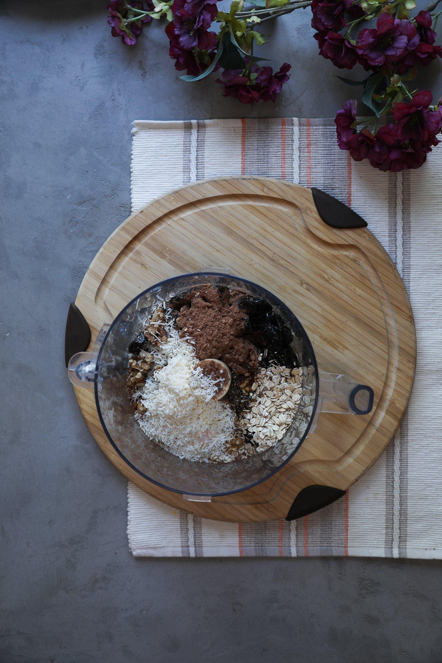 Unblended ingredients for chocolate coconut ladoos in a food processor, including oats, coconut, cocoa powder, prunes, and walnuts.