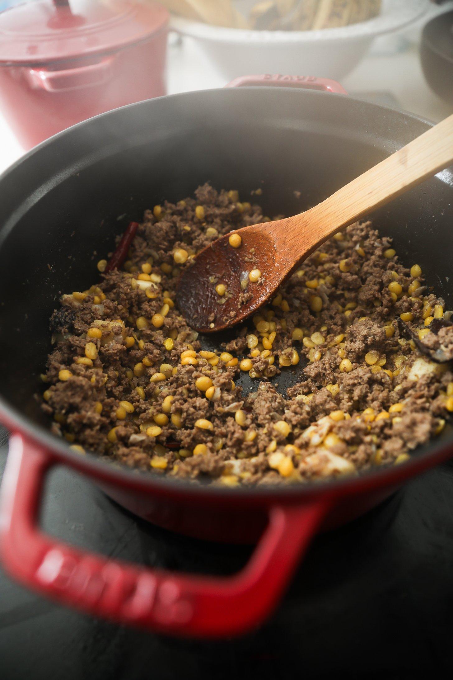 Beef and chana dal mixture cooking down in red Dutch oven with wooden spoon.