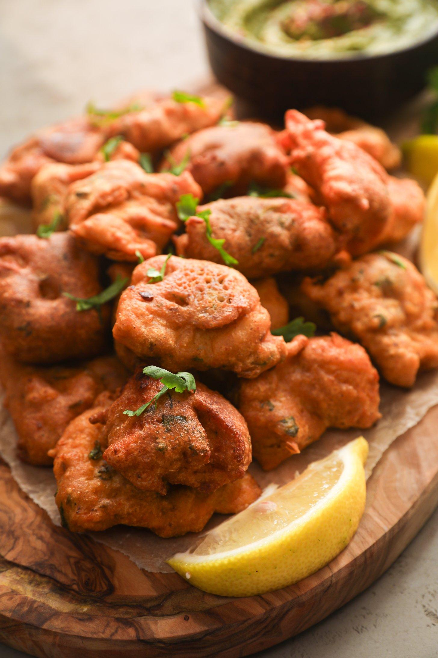 Close-up of crispy, golden-brown pakora (fritters) garnished with fresh coriander, served on a rustic wooden board with a wedge of lemon and chutney in the background.