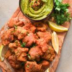 Plate of crispy basa fish pakora fritters garnished with fresh coriander leaves and lemon wedges on a rustic wooden board, accompanied by a bowl of vibrant green chutney topped with red chili flakes.
