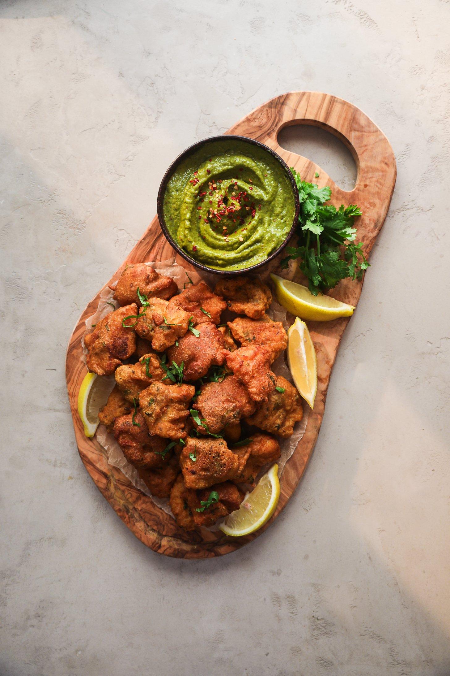 Golden crispy pakora fritters garnished with chopped cilantro, lemon wedges, and a bowl of green chutney on a wooden serving board.