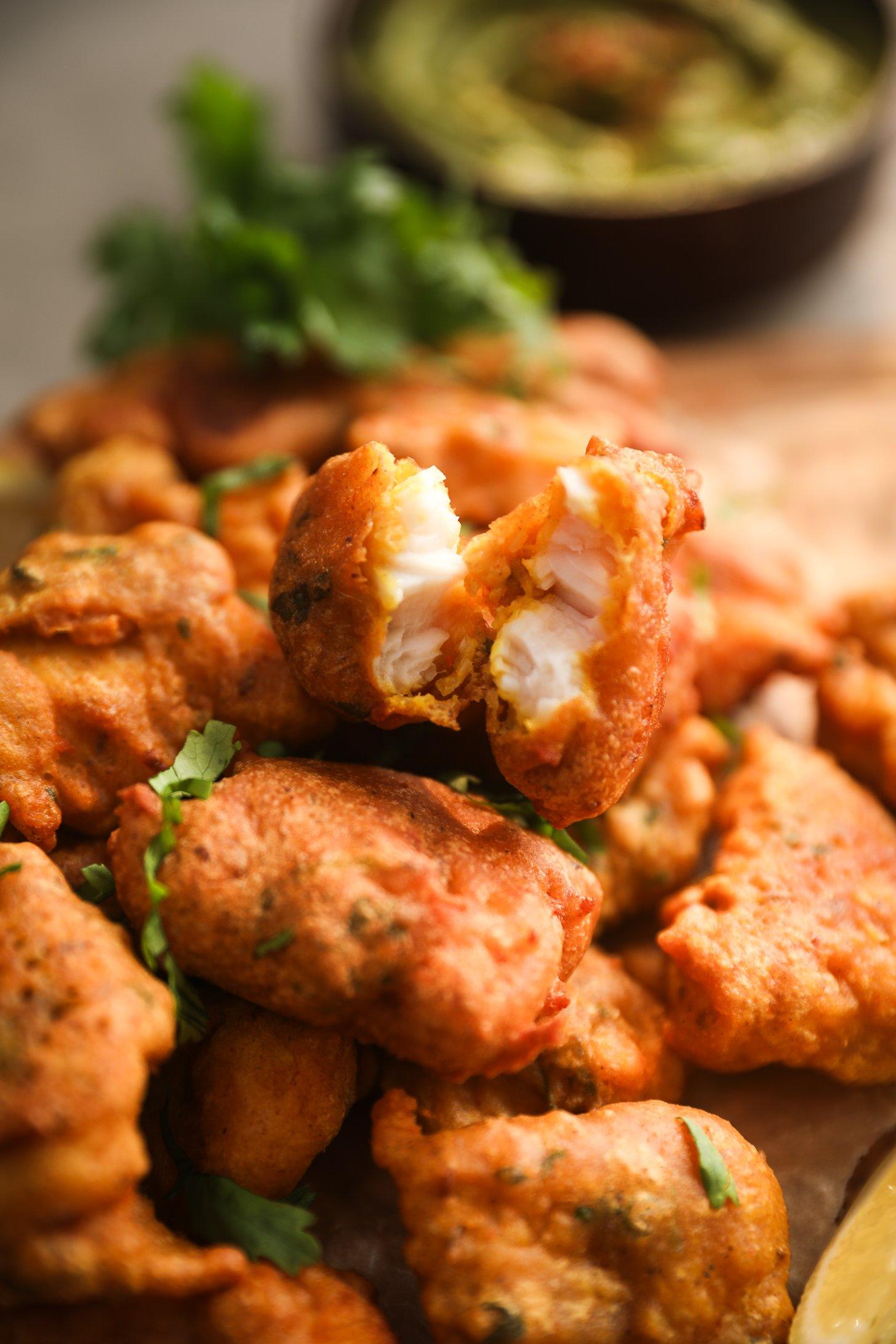 Close-up of crispy fish pakoras showing the golden-brown batter exterior with a piece broken open to reveal the flaky, moist white fish inside. Garnished with fresh cilantro.