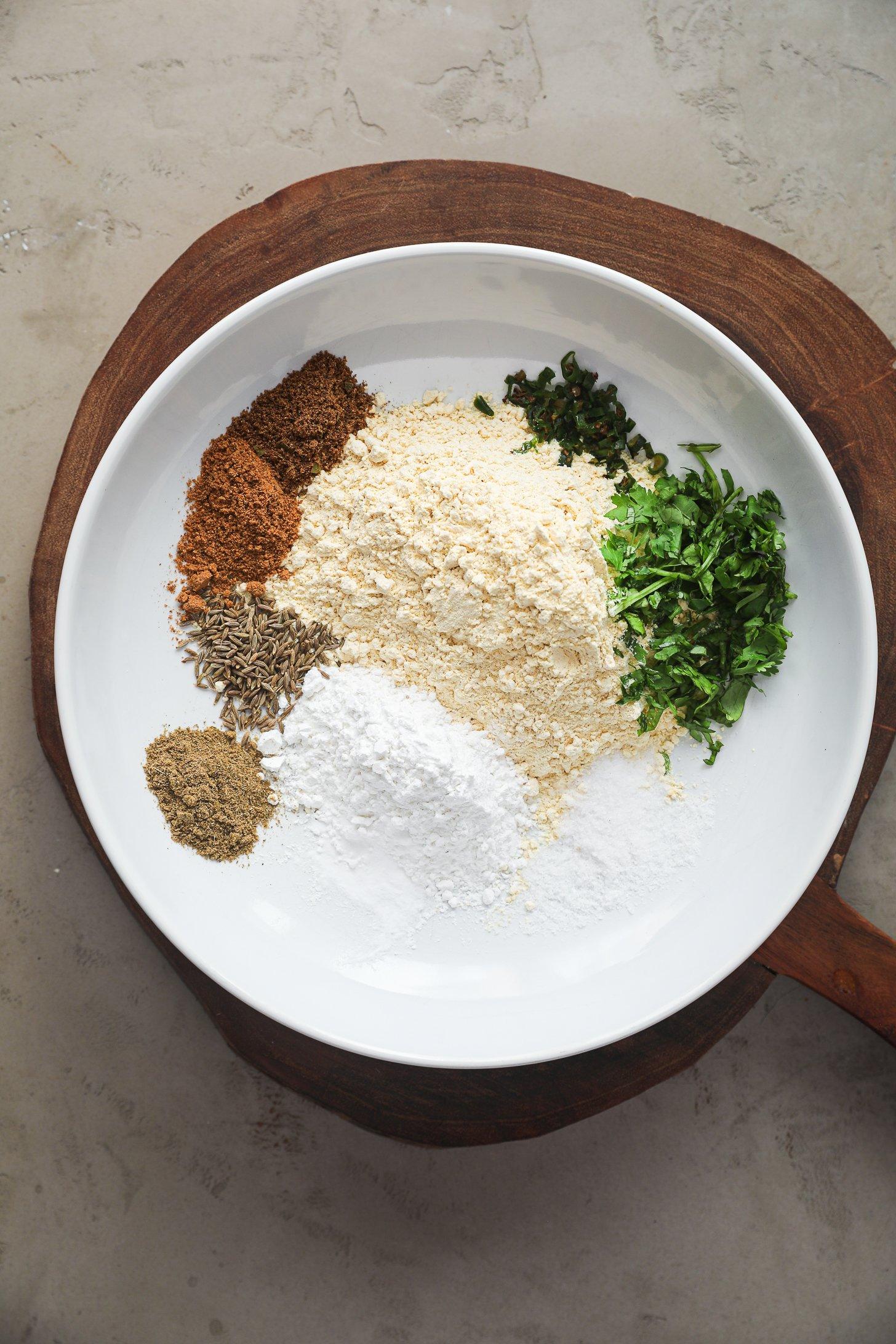 White bowl with measured spices including cumin seeds, ground coriander, garam masala, turmeric, chickpea flour, and fresh chopped green chili and cilantro.