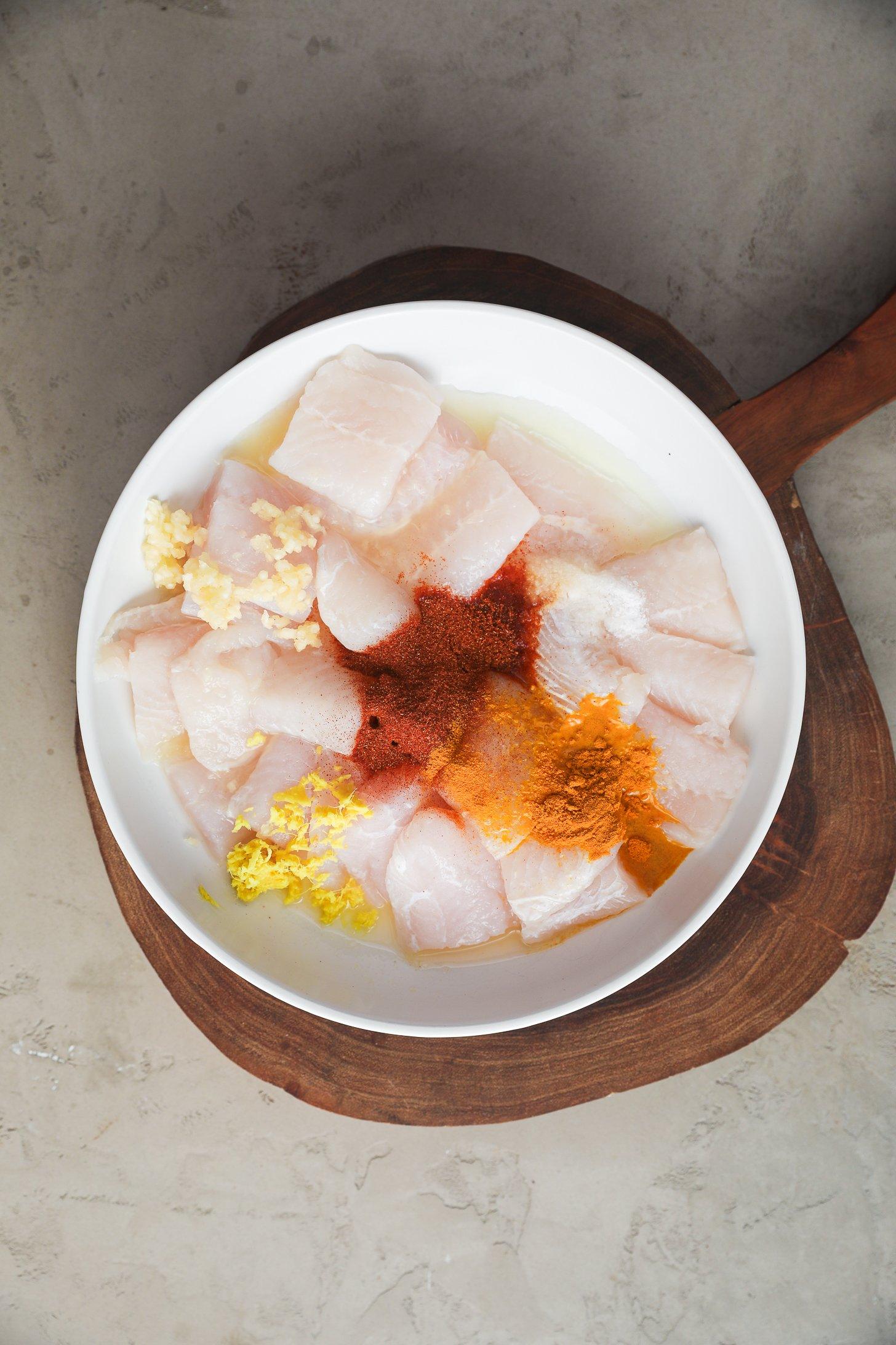 Unfried basa fish pieces in a bowl topped with powdered spices, minced garlic and ginger.