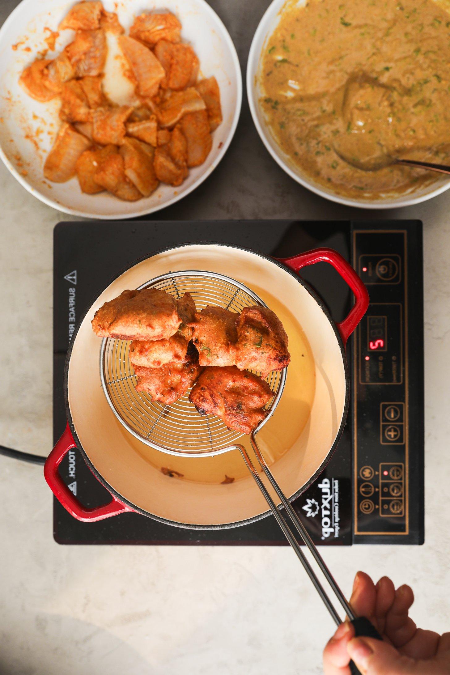 Overhead view of hand holding a slotted skimmer lifting golden brown basa fish pakoras from hot oil in a red deep pot on an induction cooktop.
