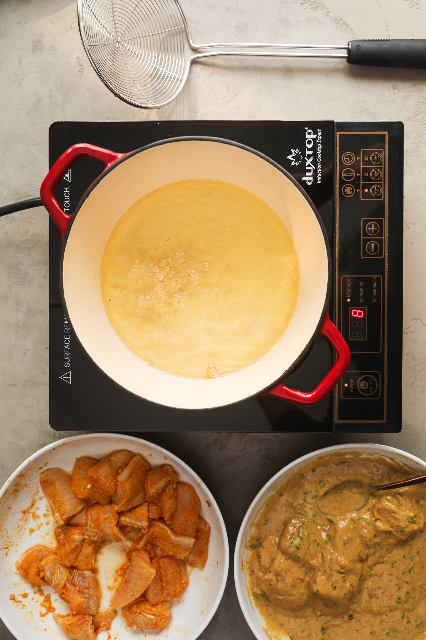 Top-down view of red enamel pot with hot oil bubbling on an induction cooktop, alongside bowls of marinated basa fish pieces and spiced chickpea batter.