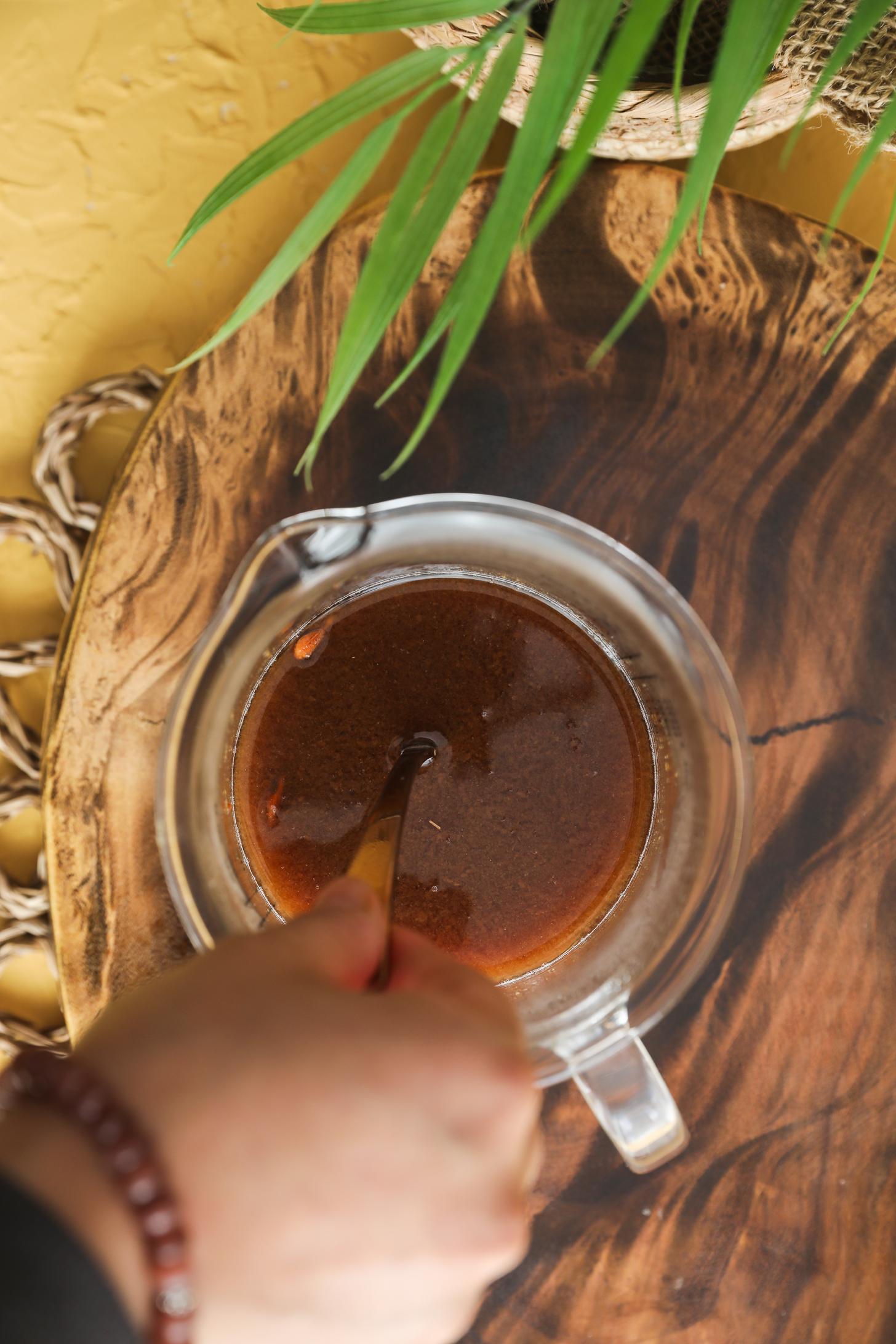 Handing holding spoon submerged inside jug of tamarind paste soaking in hot water in a clear jug.
