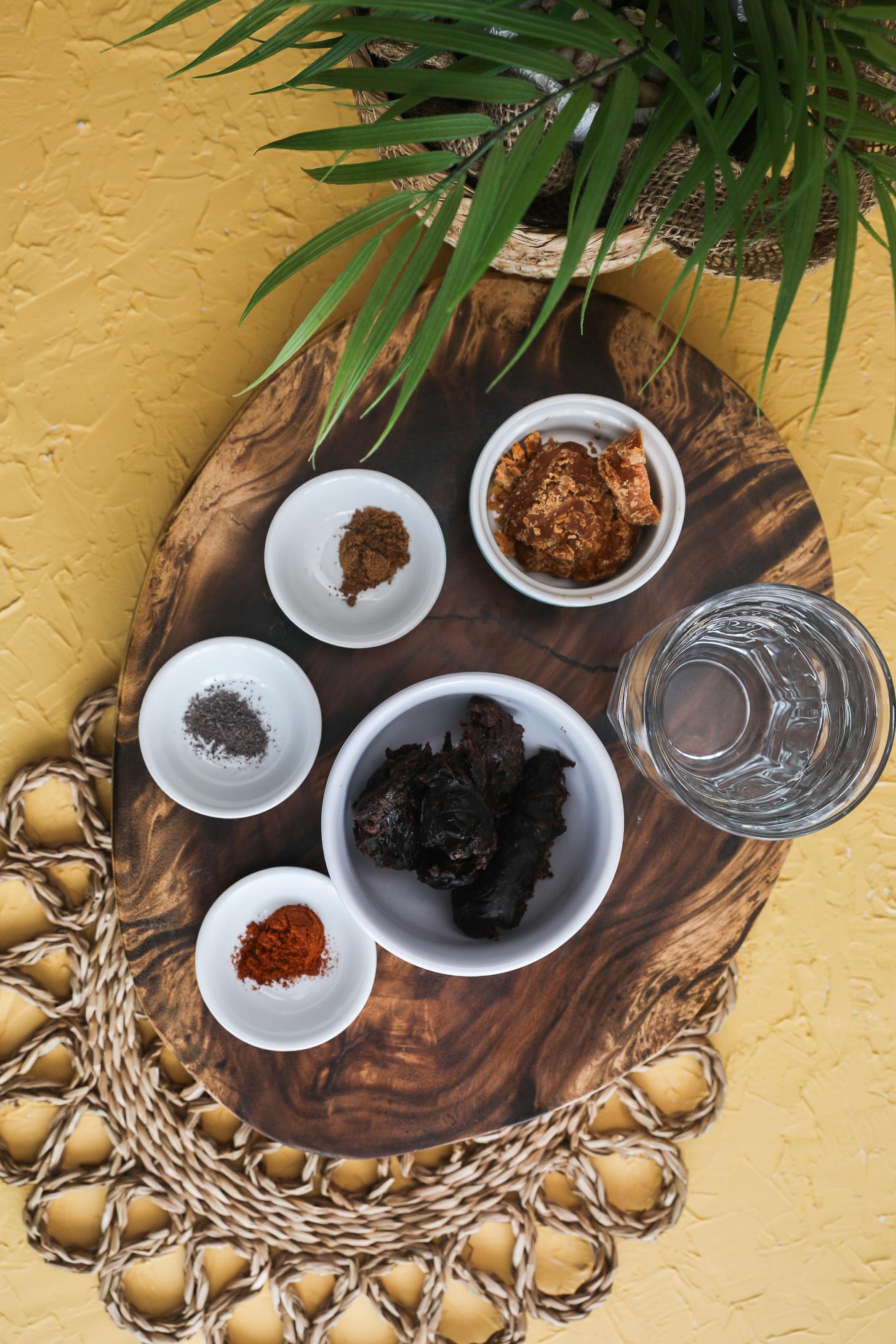 Bowls of tamarind pulp, jaggery, cumin powder, black salt, chili powder, and water on wooden board. with a green plant nearby.